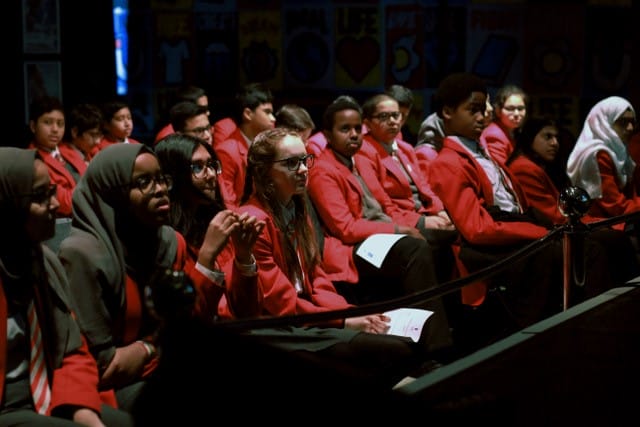 School children, dressed in uniform, are seated in a cinema screen watching a film.