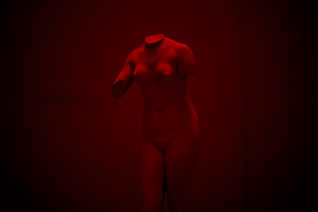 An ancient Greek statue of a naked woman, with no arms or head is light by a dark red light in this image from Vilnius Takes Over festival,