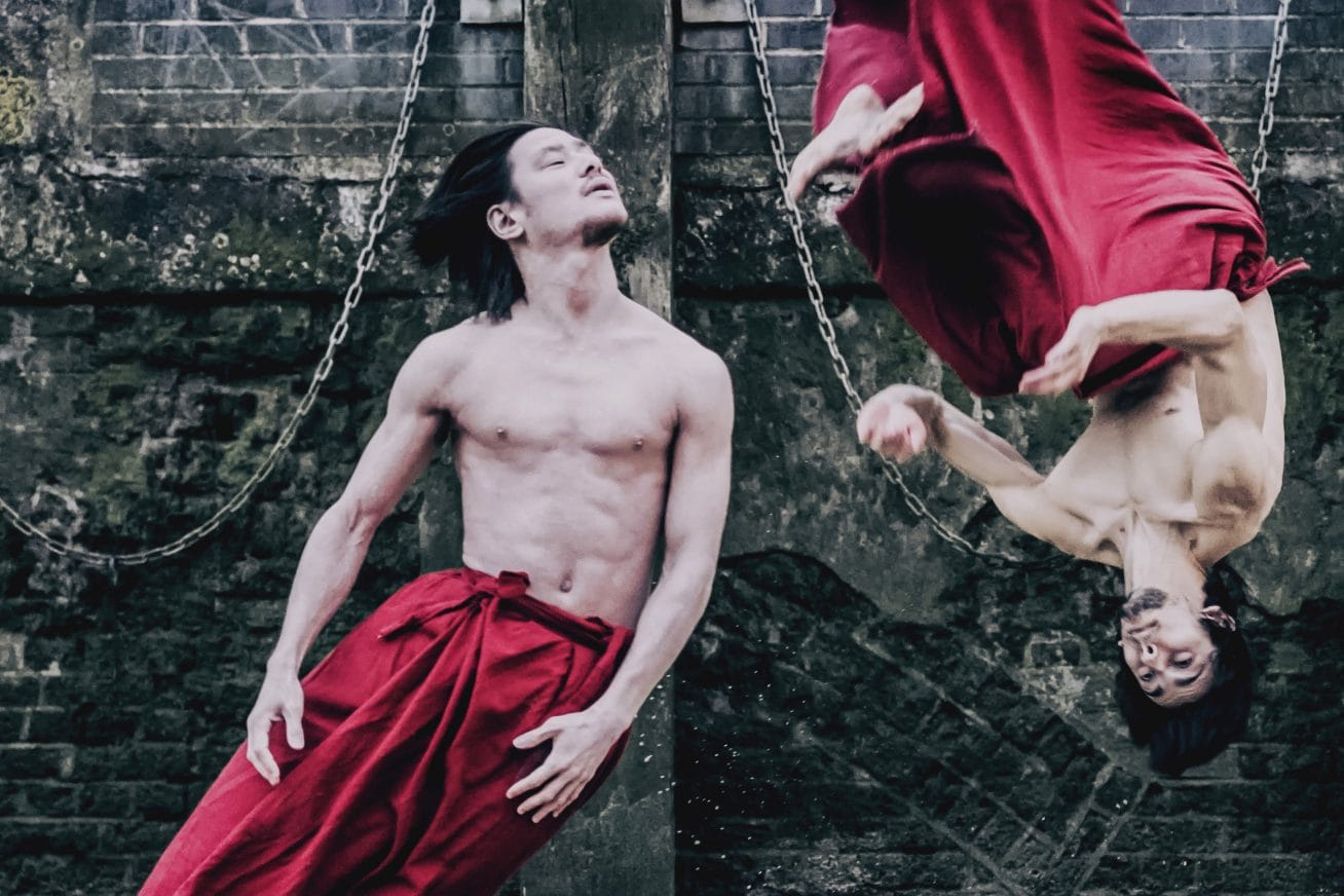 Two men in striking red sarongs take part in energetic dance moves. Their torsos are naked and muscular. The ground is sandy, and behind them sits a brick wall in this image from Chinese Arts Now Festival's Red INK.