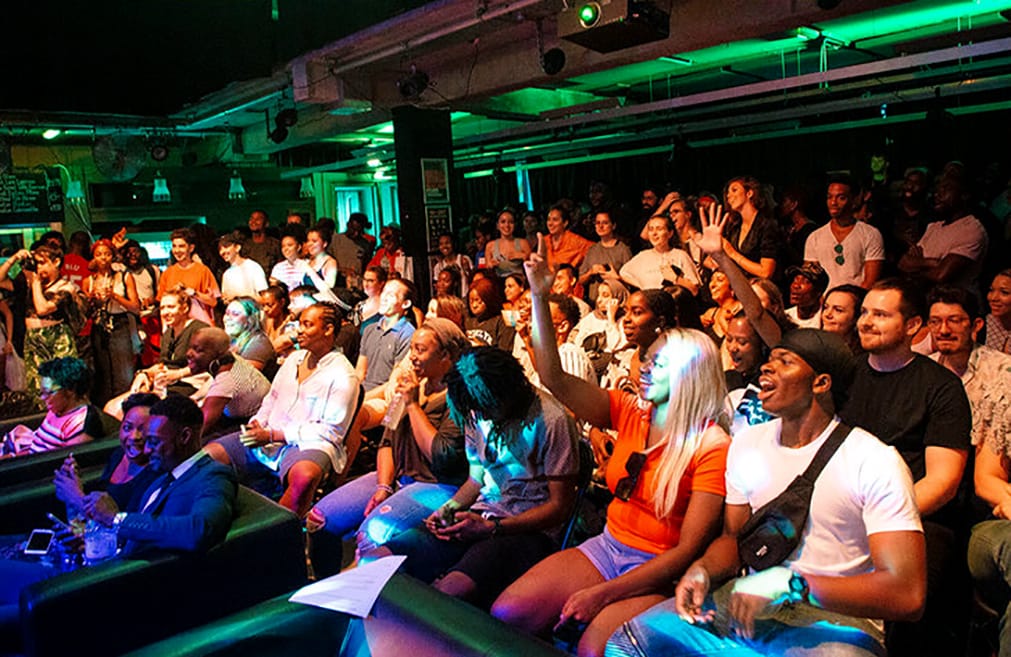 A vibrant, diverse and excited crowd waves towards a stage in front of them enthusiastically, in this image from a Bounce Cinema event at Rich Mix.
