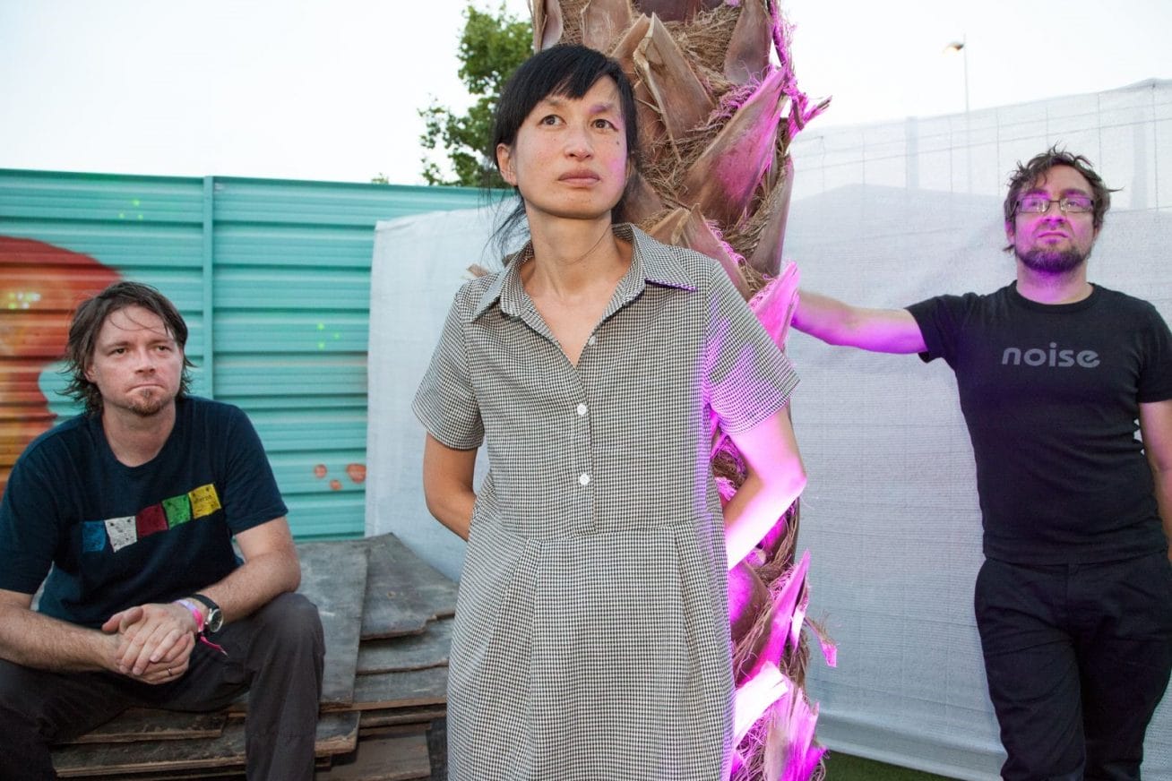 To the left a man dressed all in black sits against a bright green fence. In the middle stands a woman leaning against a palm tree with a bright pink light shining light upwards onto her arm. On the right, another man dressed all in black leans against the tree. The image is of the German elctropop band, who will do a music gig at Rich Mix.