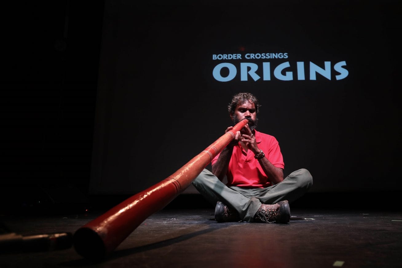 A person sits on a stage playing a didgeridoo