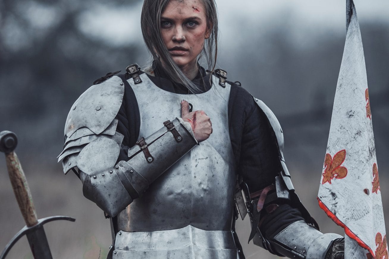 A woman wears armour as if she's in battle, and holds her fist over her heart