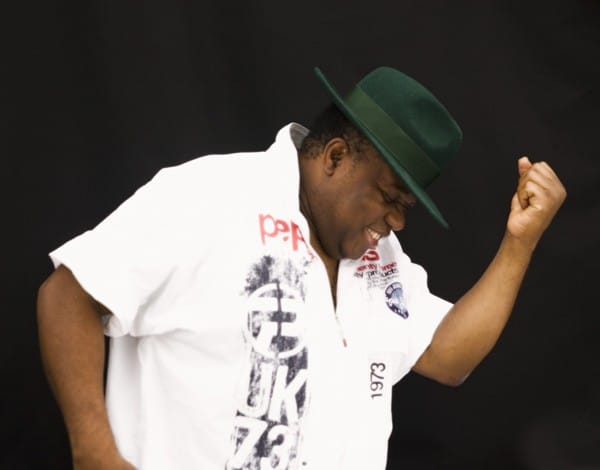 A man poses in a white shirt and fedora-style hat