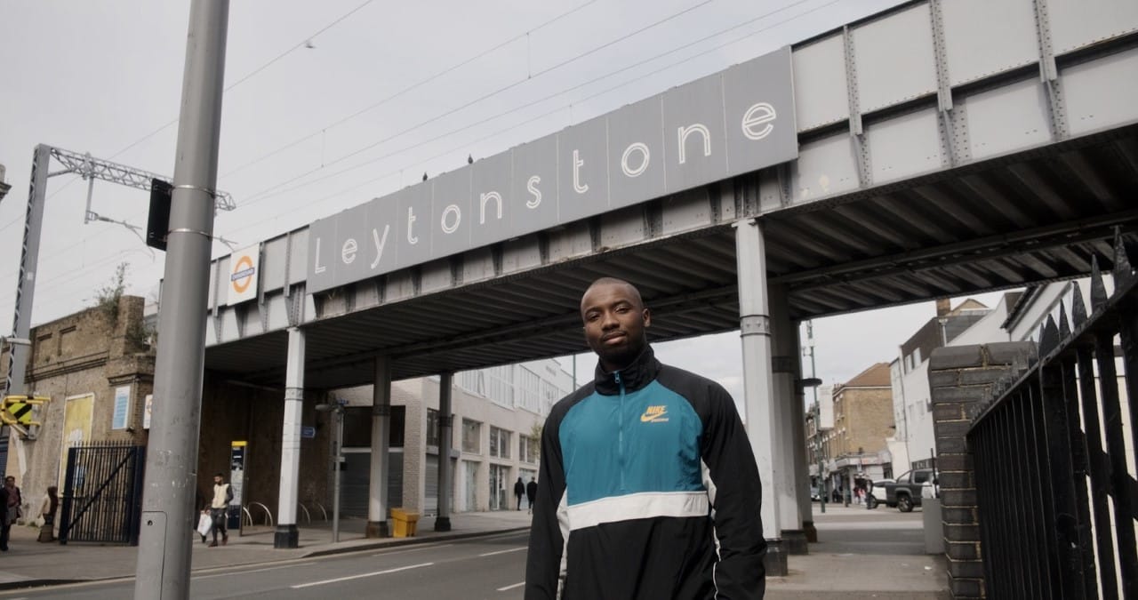 A man dressed in a blue white and black tracksuit jacket stands in front of a sign that says Leytonstone