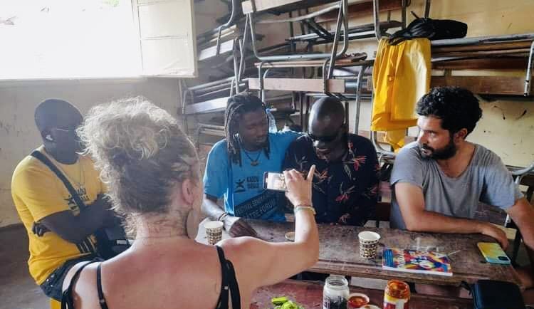 This is a sensory workshop scene, with five participants. In the foreground a white woman with blonde curly hair (Creative Director Amy Neilson Smith) holds a camera towards a black man (VI associating), with black glasses. On the table there are sensory tastes and smells, including green mint, black olives and red chilli. To the left, Associate Artist Gustavo Dias, turns to listen to the sensory conversation.